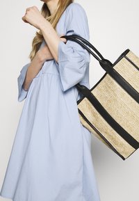 Stroh-Tote-Bag mit schwarzen Lederakzenten und -griffen, ausgestattet mit einem strukturierten, gewobenen Design. Das Modell trägt ein hellblaues, langärmliches Kleid.