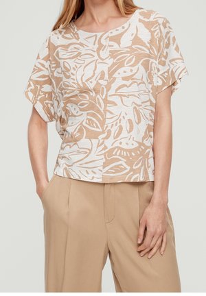 Kobieta w beżowych, szerokich spodniach i krótki rękawem bluzce w beżowo-biały abstrakcyjny wzór liści, z okrągłym dekoltem.