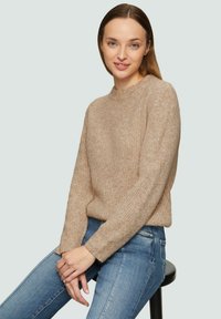Beiger Strickpullover mit einem hohen Rundhalsausschnitt und langen Ärmeln, der ein strukturiertes Muster aufweist. Kombiniert mit blauen Jeans, sitzt auf einem schwarzen Hocker.