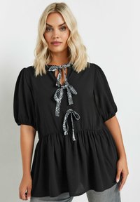 Blusa nera con maniche a sbuffo, scollo annodato e vita arricciata. Gli accessori includono nastri fantasia e un tessuto morbido e texture.