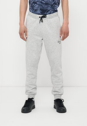 Moški v svetlo sivih trenirkah z črnim vrvico in črnimi supergami stoji pred enobarvnim belim ozadjem.