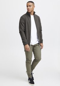 Donkergrijze zip-up jas met staande kraag, witte t-shirt eronder, olijfgroene strakke broek en zwarte sneakers met witte accenten.