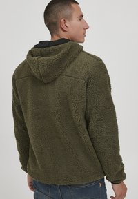 Olive green fleece jacket with a textured surface, hood, and black lining. Features a relaxed fit and side seams for added detail.