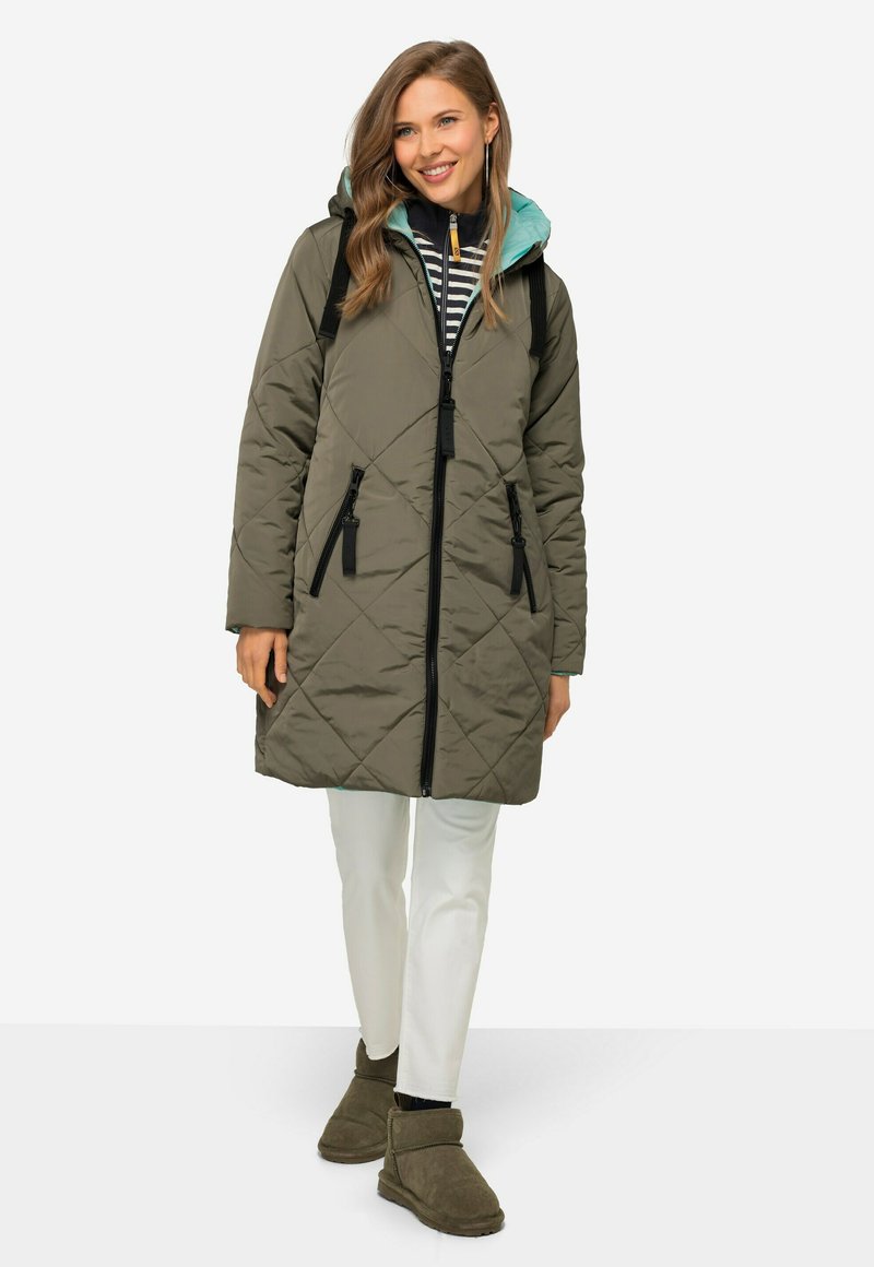 Cappotto imbottito verde oliva con motivo a rombi, chiusura con zip, dettagli neri, tasche laterali e cappuccio azzurro, abbinato a pantaloni bianchi.