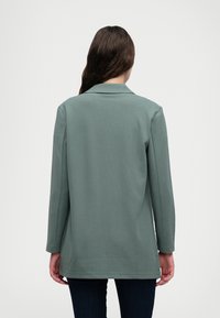 Lys grøn blazer med krage, lange ærmer og lige snit. Stoffet ser blødt ud; har minimal syning og ingen synlig hardware.