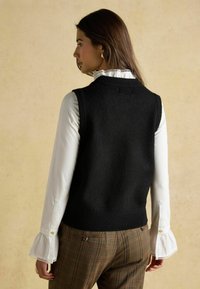 Must must vest, mille ümar kaelus, kantud valge pluusi üle, millel on rüüsosed varrukad, kombineeritud ruuduliste pükstega. Tagumine vaade, neutraalne taust.