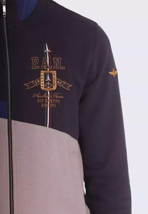 Giacca zip-up blu notte e crema con ricami dorati, caratterizzata da un logo, strisce e una finitura testurizzata. Chiusura con zip e polsini elasticizzati.