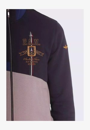 Giacca zip-up blu notte e crema con ricami dorati, caratterizzata da un logo, strisce e una finitura testurizzata. Chiusura con zip e polsini elasticizzati.