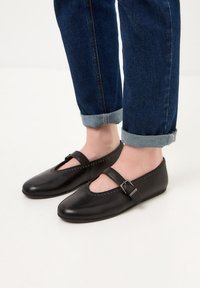 Schwarze Leder-Ballettschuhe mit einem ausgeschnittenen Design und einem seitlichen Schnallenriemen, verziert mit kleinen Nieten entlang der Kante. Getragen mit hochgekrempelten Jeans.