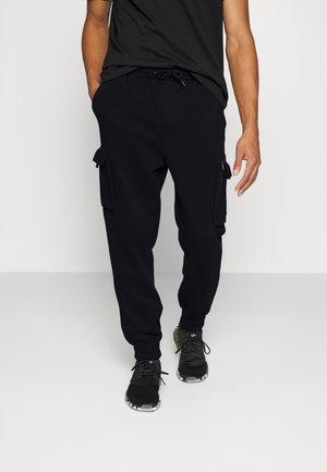 Homme portant un pantalon cargo jogger noir avec des poignets élastiques et des baskets noires, debout sur un fond blanc.