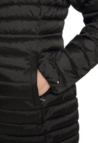 Schwarze Pufferjacke mit gestepptem Design, ausgestattet mit einer mit Reißverschluss versehenen Tasche und einem kleinen Logoakzent am Ärmel. Weiche, glänzende Textur.