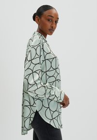 Blusa verde chiaro con motivo circolare nero, maniche lunghe, colletto e spacchi laterali. Realizzata in tessuto morbido con una vestibilità comoda.