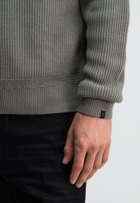 Gebreide grijze trui met ribbeltextuur, voorzien van een manchet detail en een klein label op de mouw. Draagt over zwarte broek.