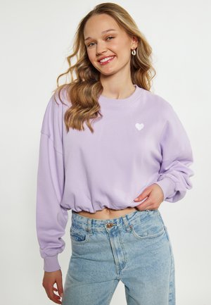 Uśmiechnięta kobieta z falującymi blond włosami nosi jasnofioletową krótką bluzę z nadrukiem serca oraz jasnoniebieskie jeansy z wysokim stanem na białym tle.