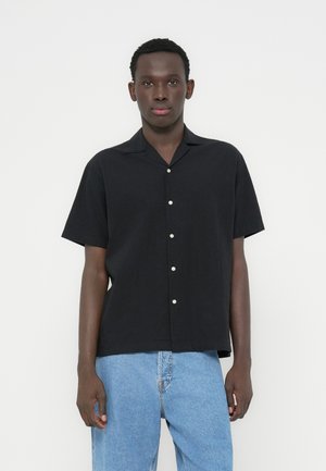 Schwarzes Kurzarmhemd aus leichtem Stoff, mit Camp-Kragen und weißen Knöpfen, kombiniert mit hellblauen Denim-Jeans.