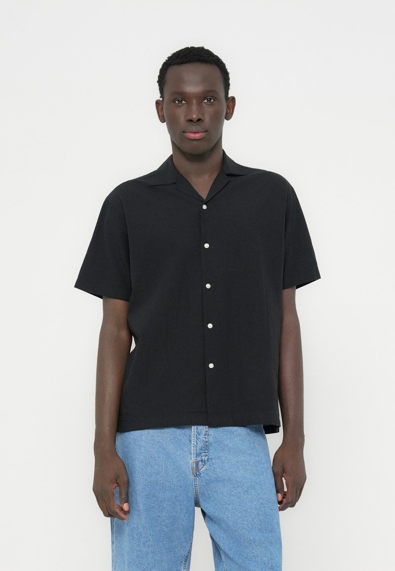 Μαύρο κοντομάνικο πουκάμισο από ελαφρύ ύφασμα, με γιακά τύπου κάμπινγκ και λευκά κουμπιά, συνδυασμένο με τζιν παντελόνι σε ανοιχτό μπλε χρώμα.
