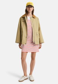 Beige jakke med brun krave, hvid kjole med røde striber og hvide sneakers. Kappen er gul med blå tekst.