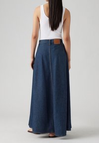 Džínová maxi sukně v tmavě modré barvě, s vysokým pasem, zapínáním na zip a koženou nášivkou na zadní straně. Hladká textura, délka až k podlaze.