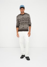 Brauner gemusterter Pullover mit weißen geometrischen Designs, kombiniert mit weißen Hosen und schwarzen Schuhen, ergänzt durch eine blaue Mütze.