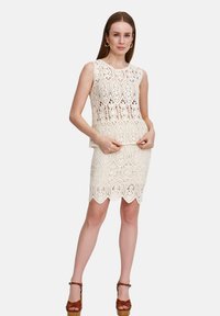 Sukienka koronkowa w kolorze białym łamanym, z falbaniastym dołem i bez rękawów, zdobiona skomplikowanymi wzorami z wycięciami. W połączeniu z brązowymi sandałami na obcasie.