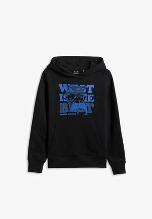 Jeune personne debout, jambes croisées, portant un sweat à capuche noir avec un graphisme bleu, un pantalon noir, des baskets et un bandeau noir à motifs sur fond gris.