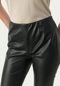 Schwarze Kunstlederhosen mit glatter Textur, hoher Taille und dezenten Nähten. Kombiniert mit einem hellbeigen Oberteil.