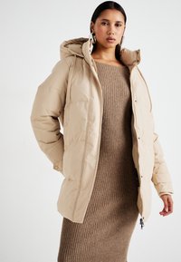 Doudoune beige à finition texturée, avec capuche et fermeture à boutons-pression, portée sur une robe marron à côtes.