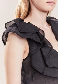 Musta hihaton toppi, jossa on röyhelökoristellut olkaimet. Kangas on kevyttä ja läpinäkyvää, luoden kerroksellisen ulkonäön.