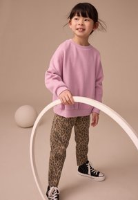 Svetloružová nadrozmerná mikina a nohavice s leopardím vzorom, doplnené čiernymi teniskami. Dieťa drží biely kruh a stojí na neutrálnom pozadí.
