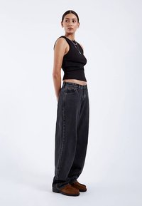 Black ribbed tank top, loose-fitting dark gray denim trousers, and brown slip-on shoes. Model stands sideways against a plain background.