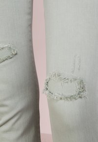 Vaaleanharmaat farkut, joissa on kuluneita kohtia, ominaisuutena revitty reunus ja sileä pinta. Lähikuva polven alueista.