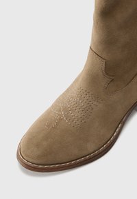 Lys brune ruskinns cowboystøvler med broderte dekorative mønstre på tåen og en mørkebrun sydd såle.