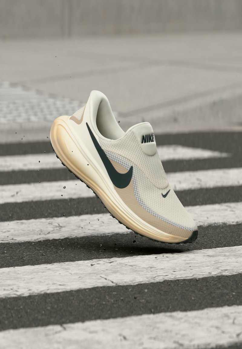 Λευκό και μπεζ αθλητικό παπούτσι Nike με μαύρο λογότυπο παύλα, στον αέρα πάνω από διάβαση πεζών σε δρόμο με άσφαλτο.