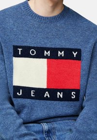 Blå stickad tröja med ett stort motiv med texten "TOMMY" och "JEANS", i röd, vit och marinblå färgblock. Rundhalsad, strukturerad tyg.