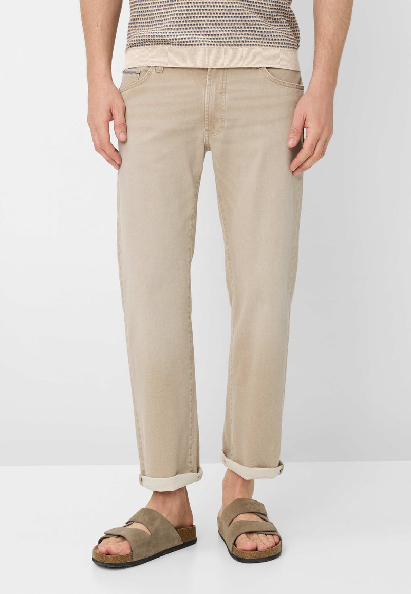 Homme portant un pantalon beige retroussé et des sandales en daim marron, debout devant un fond blanc.