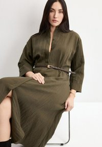 Žena s dlouhými tmavými vlasy sedící na kovové židli, oblečená v zapnutých olivově zelených šatech s tenkým tmavým páskem a černými botami.