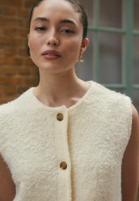 Gilet sans manches blanc texturé avec un col arrondi, doté de deux boutons marron sur le devant et d'une finition en tissu doux et pelucheux.