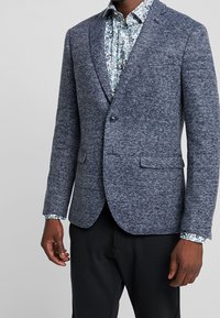 Marinblå ullblandad kavaj med texturerad finish, enkel knappstängning och två framfickor. Under den syns en mönstrad skjorta.