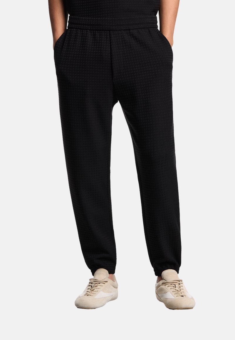 Joggers neri con motivo, cintura elastica, tasche laterali e gambe affusolate, abbinati a scarpe da ginnastica di colore chiaro.