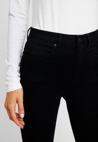 Femme portant un jean noir taille haute avec poches avant et une chemise blanche à manches longues, la main reposant près de la cuisse.