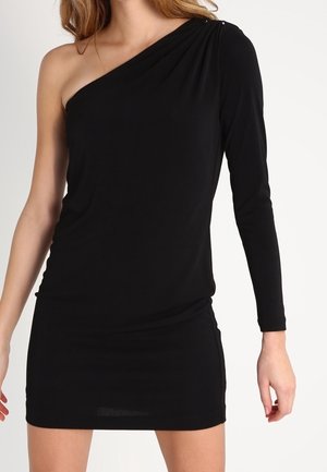 Frau trägt ein schwarzes Mini-Kleid mit einem Schulterriemen und einem langen Ärmel am rechten Arm vor einfarbigem Hintergrund.