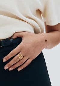 Kullanılan bir elde, dokulu dikenler ve geometrik şekillerle süslenmiş altın bir yüzük ile üzerinde bir 'S' harfi bulunan zarif bir altın bilezik.