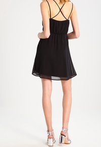 Robe sans manches noire avec une taille ajustée et une jupe évasée, dotée de bretelles croisées dans le dos. Porte des sandales à talons hauts argentées et brillantes.