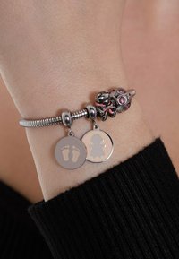 Silberarmband mit einem flexiblen Design, ausgestattet mit einem Anhänger in Pink und zwei Anhängern mit einem Babyfußabdruck und einer Silhouette.