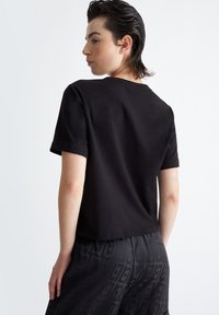 T-shirt nera a maniche corte, in materiale cotone, vestibilità comoda, scollatura a girocollo, con maniche arrotolate, indossata con pantaloni corti neri a fantasia.