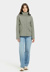 Grøn, vandtæt jakke med høj krave og hætte, som har lynlåslommer på siden, kombineret med lyseblå jeans og beige sneakers.