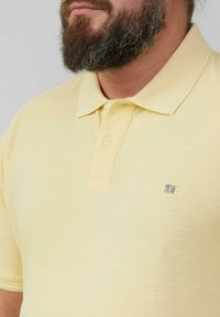 Gelbes Polohemd aus strukturiertem Stoff, klassischem Kragen und drei Knöpfen, mit einem kleinen Logo auf der linken Brust.