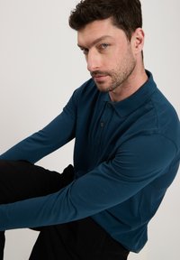 Koszulka polo z d ługim rękawem, turkusowa, wykonana z miękkiego materiału, z kołnierzykiem i zapięciem na trzy guziki, z logo na klatce piersiowej.