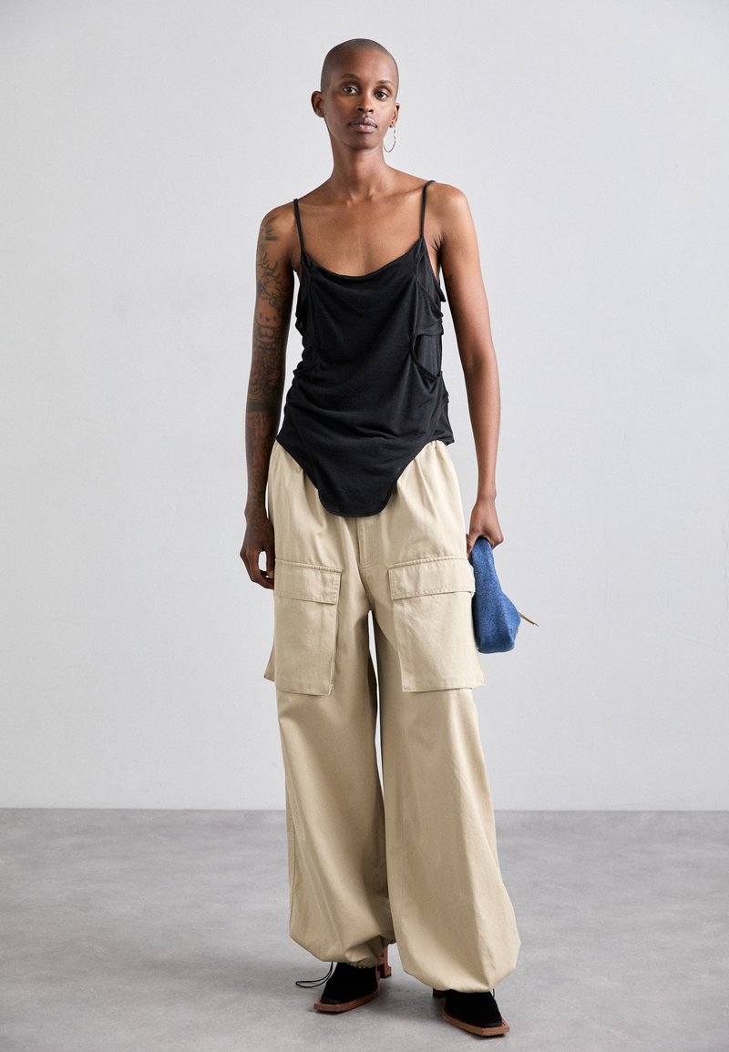 Schwarzes, leichtes Tanktop mit Drapierung, kombiniert mit cremefarbenen Cargo-Hosen mit großen Taschen und schwarzen Schuhen; blauer Beutel in der Hand.