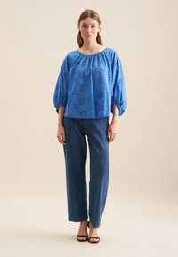 Blå bomuldsbluse med øjelet design, brede ærmer og rund halsudskæring, parret med højtaljede straight-leg jeans og sorte sandaler.